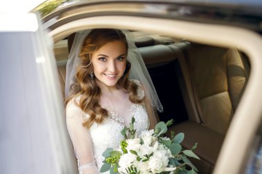 Düğününde gülümseyen güzel bir gelin arabada oturur ve elinde bir buket beyaz gül tutar. Güzel saç stili olan genç bir kadın kendi düğününde sevinir.