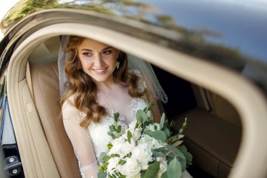 Genç, gülümseyen bir gelin pahalı bir arabada oturur ve elinde beyaz bir buket gül tutar. Mutlu gelin kendi düğün gününün tadını çıkarıyor. Metin için boş alan. Gelin için güzel saç stili.