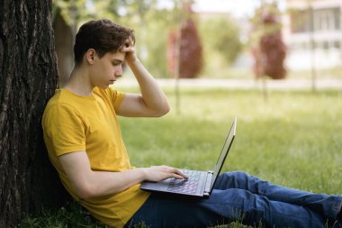 Sarı tişörtlü genç bir adam çimlerde oturur ve dizüstü bilgisayarla sıkı çalışır. Modern teknolojiler uzaktan çalışmanızı sağlar. Serbest çalışan biri dışarıda çalışıyor. Metin için boş alan