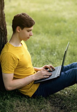 Genç bir adam çimenlerde oturuyor ve elinde bir dizüstü bilgisayar tutuyor. Okuldan sonra dinlenmek ya da açık havada öğretmenlik yapmak. Modern teknolojileri kullanarak uzaktan çalışma. Serbest çalışan birinin dikey fotoğrafı.
