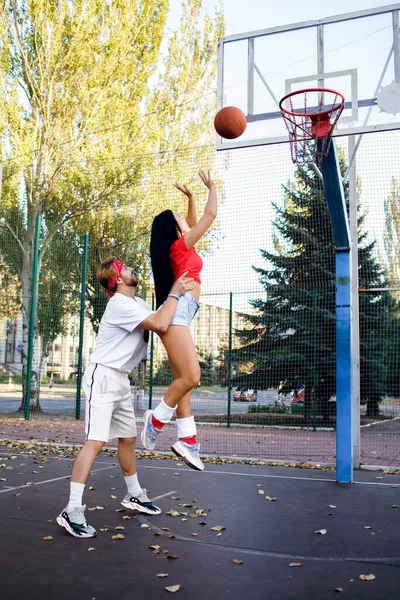 Young sportive couple playing basketball. A beautiful girl and a sporty guy go in for sports in their free time. Heterosexual couple in love loves sports. The guy puts the girl on the basketball hoop