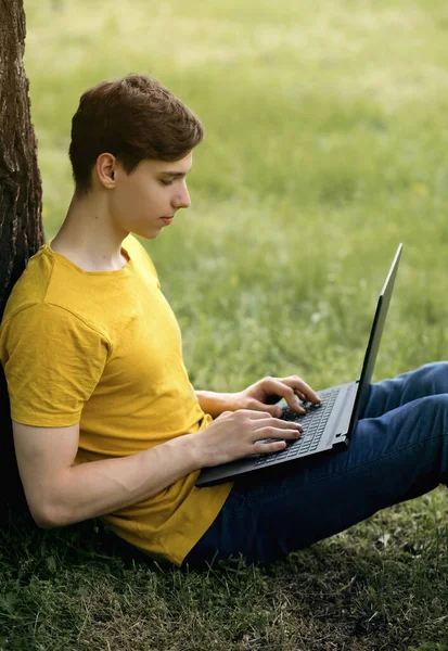 A young guy sits on the lawn and holds a laptop in his hands. Relax after school or teach remotely in the great outdoors. Remote work using modern technologies. Vertical photo of a freelance worker