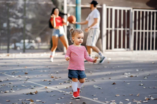 An athletic family spends the evening on the basketball court playing sports. A little girl holds a dry leaf while Mom and Dad play basketball. Happy young family playing sports on weekend