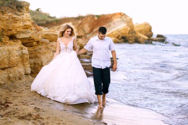Güzel genç gelin ve damat deniz boyunca kumlu kayalıklarda yürüyor. Yakışıklı damat, beyaz gelinlik giymiş zarif bir gelinin elini tutuyor. Mutlu yeni evliler deniz kıyısında yürür