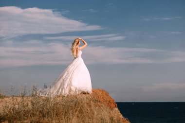 Uzun beyaz gelinlik içinde güzel bir gelin dağın kenarına doğru yürür. Genç, gülümseyen bir gelin bir dağın kenarında durur ve okyanusa bakar. Parlak renkli düğün fotoğrafı.