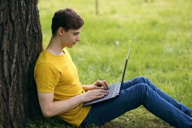 Genç bir adam çimenlerde oturuyor ve elinde bir dizüstü bilgisayar tutuyor. Okuldan sonra dinlenin ya da dışarıda çalışın. Modern teknolojileri kullanarak uzaktan çalışma. Rahatlık içinde serbest çalışmak. Metin için boş alan