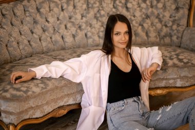 Güzel genç bir esmer dirsekleri kanepede dinlenirken yerde oturuyor. Kotlu ve beyaz gömlekli tatlı bir kız seksi görünüyor. Evde poz veren şık bir genç kadın.