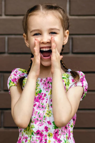 Baby girl screams loudly, holding her hands to her mouth. A preschool girl stands by a brick wall and calls out loudly to someone. Free space for text. Bright clothes and stylish hairstyle