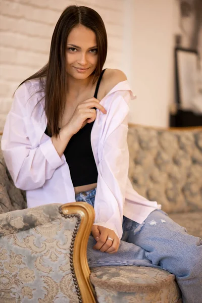 美しいセクシーな女の子モデルがソファに座って白いシャツを脱いでいます 若い美しいブルネットが魅惑的かつ反抗的に見えます 自宅でソファに座って官能的なモデルの肖像 — ストック写真