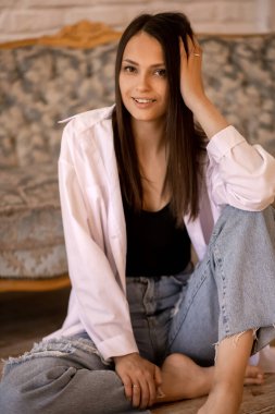 Güzel genç bir esmer yerde oturuyor, dirseklerini kanepeye dayıyor ve gülümsüyor. Kotlu ve beyaz gömlekli tatlı bir kız uzun saçlarını düzeltiyor. Seksi genç kadın delici bir şekilde bakıyor.