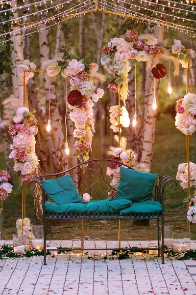 A magical decor of fresh flowers of feathers and garlands with a decorative bench. A beautiful decorated place in the evening park with candles. Bright vertical photo for social networks