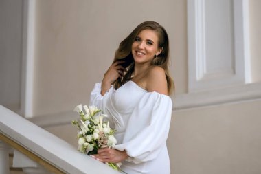 Beyaz gelinlik içinde, elinde bir buket beyaz çiçekle güzel, neşeli bir gelin. Genç bir kadın evleniyor. Zarif saç stili ve makyajlı şık bir gelin portresi.