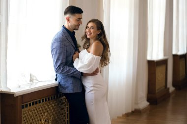 Gelin ve damat pencerenin yanında sarılıyorlar. Gelin özel yapım beyaz bir gelinlik giyiyor. Damat sevgilisine bakar ve gülümser. İçeride yeni evlilerin portresi.