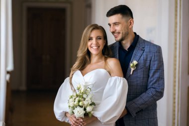 Güzel bir gelin ve damadın portresi. Mutlu gelin gülümsüyor ve elinde bir buket düğün çiçeği tutuyor. Zarif damat, sevdiği kadını sevinçle kucaklıyor ve kucaklıyor. Mutlu yeni evliler düğün günlerinde