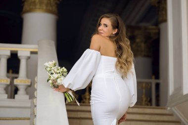 Şık beyaz elbiseli mutlu bir kız ve elinde güzel bir buket çiçekle merdivenlerden eve çıkar ve cilveli bir şekilde etrafına bakar. Zarif saç stili ve gülümseyen güzel gözleri olan genç bir kadın.