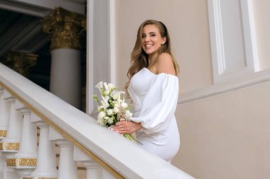 Beyaz tasarımcı elbisesi içinde bir buket çiçekli mutlu genç gelin evin içinde duruyor. Zarif bir kız düğün gününde gülümsüyor ve evlenmeye hazırlanıyor.