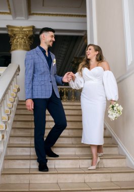 Neşeli gelin ve damat el ele tutuşarak merdivenlerden iniyor. Mutlu yeni evliler düğün günlerinde güler ve sevinirler. Gelin, şık bir gelinlik içinde beyaz bir buket tutuyor.