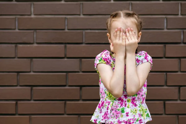 Çocuk kız avuçlarını tuğla bir duvarın arkasına koyup yüzünü kapatıyor. At kuyruklu küçük kız elleriyle gözlerini kapatıyor. Yazmak için boş yeri olan duygusal bir çocuğun portresi