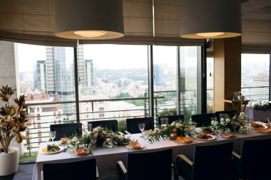 Restorandaki bir masaya şehre bakan büyük bir pencerenin arka planı servis edilir. Kafe salonu şenlikli bir akşam yemeği için misafirleri bekliyor. Festival masası çiçeklerle süslenmiş.
