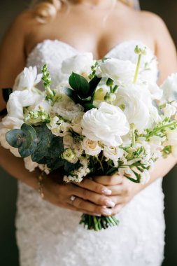Zarif elbiseli gelin elinde bir buket narin beyaz çiçek tutuyor. Çeşitli beyaz çiçekleri ve okaliptüsleri olan güzel buket gelinin ellerinde.