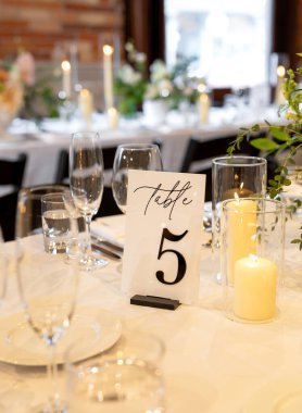 Masa 5 işareti, mumlar ve beyaz masa örtüsünün üzerindeki cam bezle şık bir düğün masası. Romantik, güzel bir yemek atmosferi var. Arkasında da sıcak mum ışığı ve çiçek dekoru var.