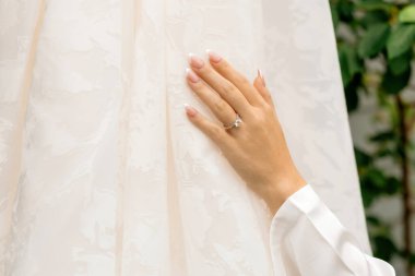 Gelinin elini nazikçe gelinliğinin üzerinde dinlendirirken, pırıl pırıl pırıl pırıl elmas bir nişan yüzüğü ve zarif bir Fransız manikürü sergiliyor - aşkın, zerafetin ve ebedi güzelliğin sembolü.