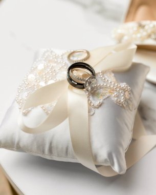 İnci, dantel ve yumuşak bir fildişi kurdeleyle süslenmiş saten bir yastıkta sergilenen alyansların zarif bir şekilde kapatılması romantik düğün ayrıntılarını ve zamansız düğün sembollerini yakalıyor.