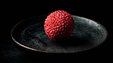 Dikenli desenli tek bir kırmızı liçi meyvesi koyu seramik bir tabakta duruyor. Dramatik ışıklandırma onun canlı rengini ve karmaşık yüzeyini vurgular, ince bir görünüm yaratır.