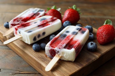 Taze böğürtlen ve nane yapraklarıyla çevrili ahşap bir tahtaya yerleştirilmiş çilek ve yaban mersini katmanları içeren canlı donmuş yoğurt dondurmaları.