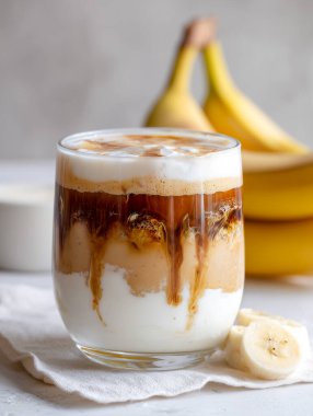 Bir bardakta kahve, süt, karamel ve fıstık ezmesi içeren katmanlı içecek, hafif bir yüzeyde taze muzlar eşliğinde.