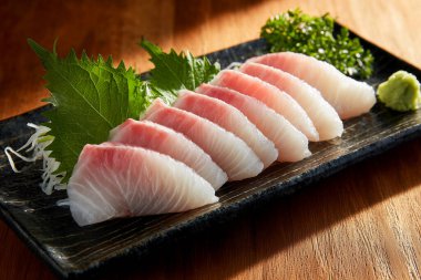 Siyah bir tabakta sergilenen taze sashimi dilimleri vasabi ve shiso yapraklarıyla birlikte, yemeğin kalitesini ve sunumunu vurguluyor.