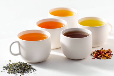 Beş beyaz fincanda bitkisel, siyah, yeşil ve meyve karışımları da dahil olmak üzere çeşitli çay türleri sergilenmekte ve beyaz bir yüzeye yayılmış çay yaprakları bulunmaktadır.