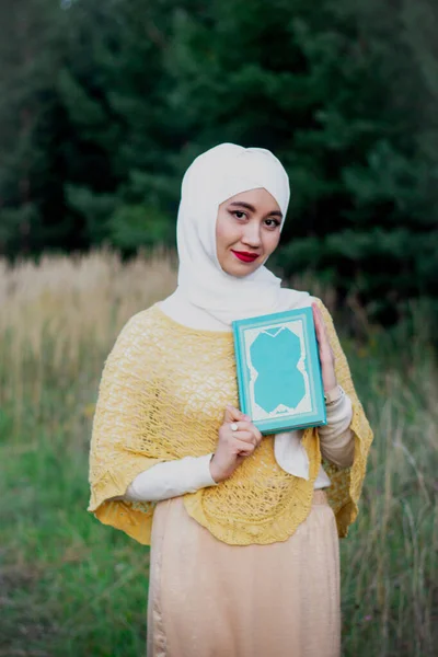 Muslim woman in hijab with a book in her hands on the field. Field near the forest. Green Book. White shawl