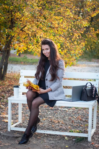Esmer bir kadın sonbahar parkında dizüstü bilgisayarı ve bir fincan kahveyle oturuyor. Öğrenci. Hocam. Çevrimiçi eğitim.