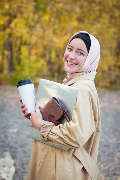 ベージュのトレンチコートを着たヒジャーブを着たイスラム教徒の女性が公園にコーヒーとノートパソコンを持って立っています ビジネスノートパッド ムスリムビジネス女性 — ストック写真