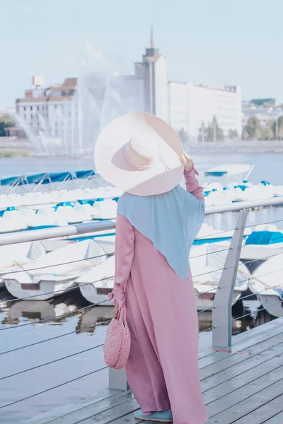 İskelede pembe tesettürlü Müslüman kadın. Hasır şapka ve pembe çanta. Yatlar arka planda.