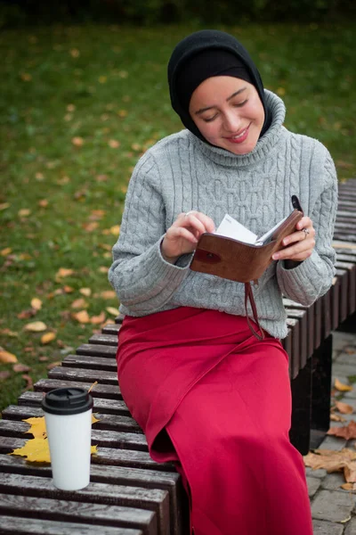 グレーのセーターを着た若いイスラム教徒の女性がベンチにノートを置いて座っています コーヒーと秋の葉のカップ 学生だ 大学生 — ストック写真