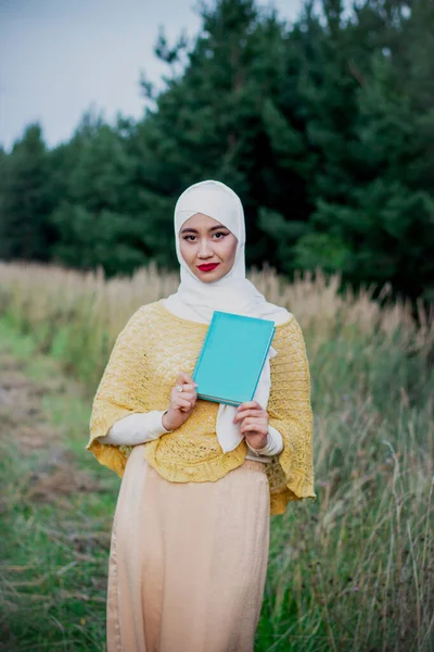 Muslim woman in hijab with a book in her hands on the field. Field near the forest. Green Book. White shawl