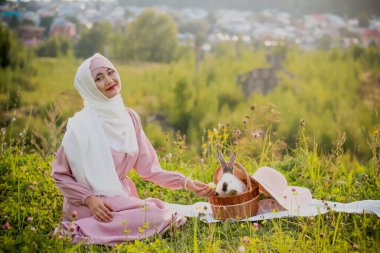 Müslüman bir kadın, elinde tavşan ve şapkayla sepetin yanında oturuyor. Pembe tesettürlü, inci bilezik takmış Müslüman bir kadının portresi. Şefkat