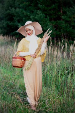 Şapkalı Müslüman bir kadın çayırda yürür ve elinde bir sepet ve kazık tutarak yürür. Bej tesettürlü Müslüman kadın. Güzellik, kadınlık, şefkat..