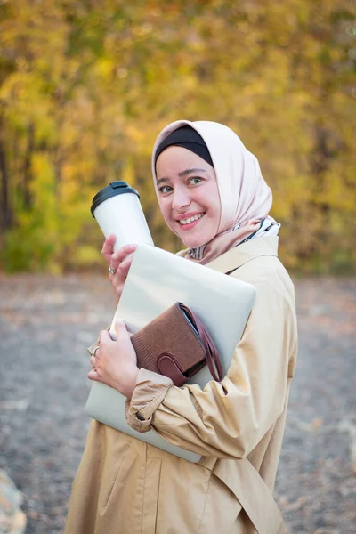 ベージュのトレンチコートを着たヒジャーブを着たイスラム教徒の女性が公園にコーヒーとノートパソコンを持って立っています ビジネスノートパッド ムスリムビジネス女性 — ストック写真