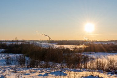 Günbatımı ve çok renkli gökyüzü ile kış akşamı manzarası. Gün batımında aydınlatılan hayvan ayak izleri olan renkli kar. Ufukta tüten bacaları olan bir sanayi bölgesi var.