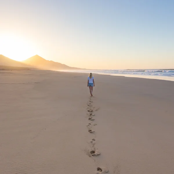 Lady walking on sandy beach in sunset leaving footprints behind. — Stock Photo, Image