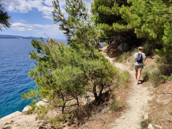 Joven turista activo con una pequeña mochila caminando por el sendero costero entre pinos que buscan una cala remota para nadar solo en paz en la costa de Croacia. Concepto de viaje y aventura — Foto de Stock