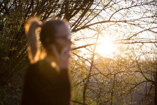 Hintergrundbeleuchtete defokussierte Rückansicht einer jungen Frau, die bei Sonnenuntergang im Park mit dem Handy telefoniert. Mädchen mit Mobiltelefon, digitalem Gerät, Blick auf die untergehende Sonne — Stockfoto