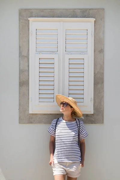 Vacker ung kvinnlig turist kvinna bär sol hatt, stående och avkopplande framför vinatage trä fönster i gamla Medelhavet stad medan sightseeing på varm sommardag — Stockfoto