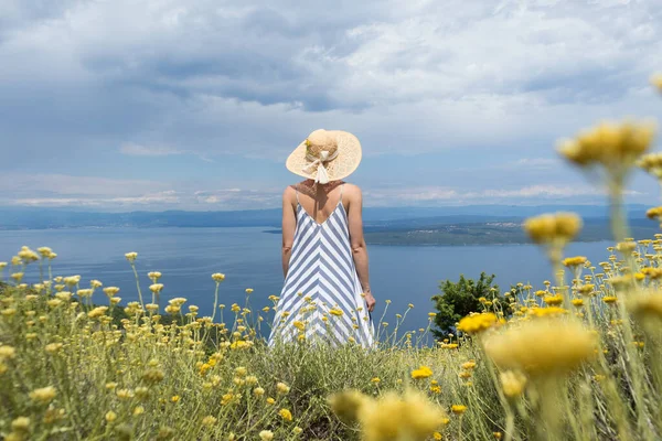 Rear view of young woman wearing striped summer dress and straw hat standing in super bloom of wildflowers, relaxing while enjoing beautiful view of Adriatic sea nature, Croatia