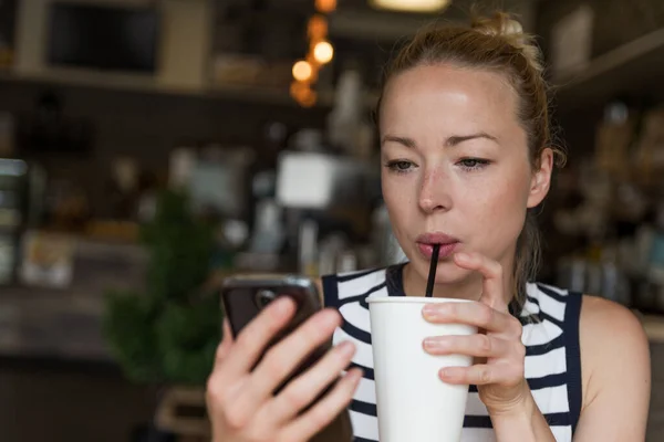 Tankeväckande kvinna läser nyheter på mobiltelefon samtidigt smuttar kaffe i caféet. — Stockfoto