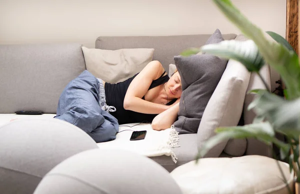 Mujer joven cansada tomando una siesta en casa acostada en un cómodo sofá con grandes cojines en la sala de estar con un teléfono móvil acostado junto a ella. — Foto de Stock