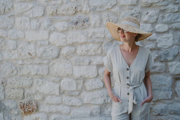 Retrato de una hermosa mujer rubia alegre con un vestido de verano de una pieza y un sombrero de verano, de pie frente a la antigua pared de piedra medieval. concepto de retrato de vacaciones de verano. —  Fotos de Stock
